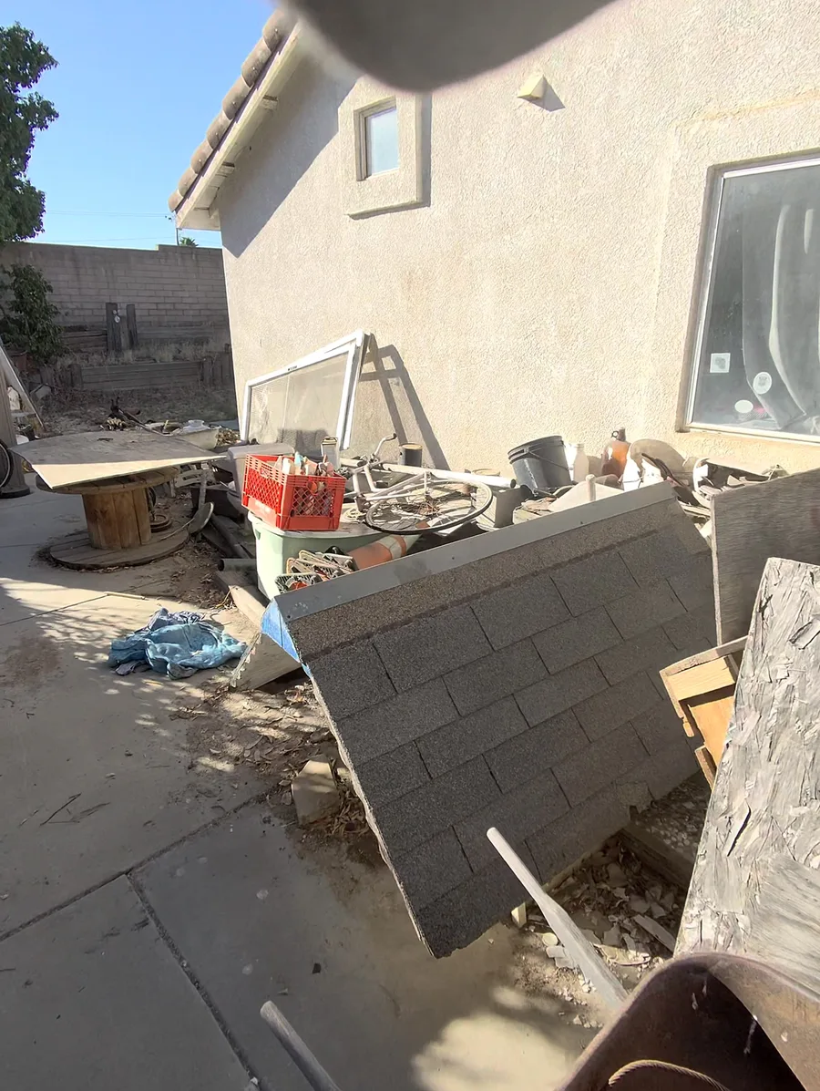 Side yard in Lake Elsinore piled with roof shingles, a scrap bike wheel, crates, and an old window frame before 1775 Junk Hauling cleared it